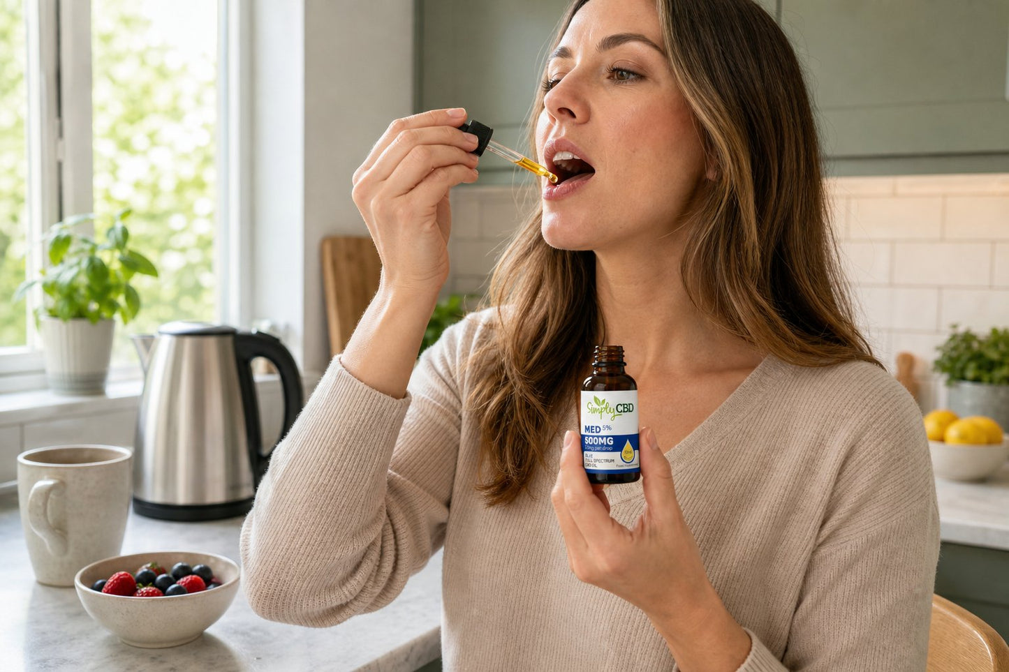 Woman in a kitchen holding a dropper to her mouth and a small bottle of liquid, with a mug, bowl of berries, kettle, and window plants on the counter nearby.